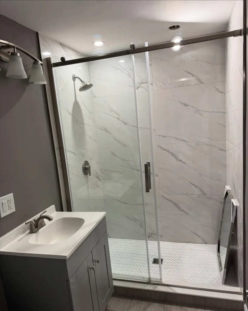 View of a completed custom tile bathroom remodel featuring a glass-enclosed shower with marble-patterned walls and a grey vanity cabinet.