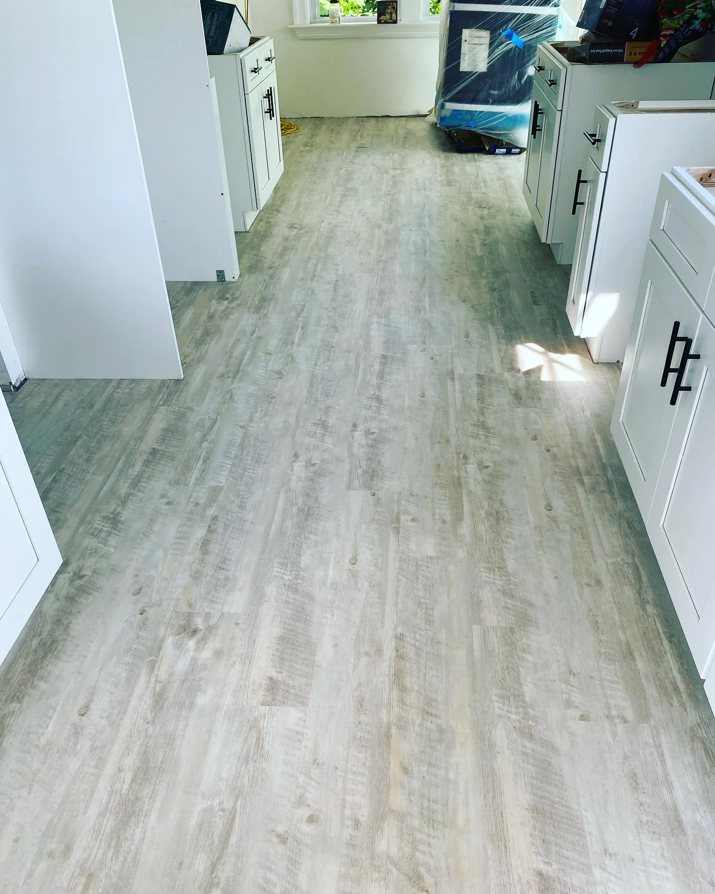 A wide perspective view looking down a hallway into a newly renovated kitchen, featuring extensive installation of modern light gray plank flooring alongside new white cabinets.