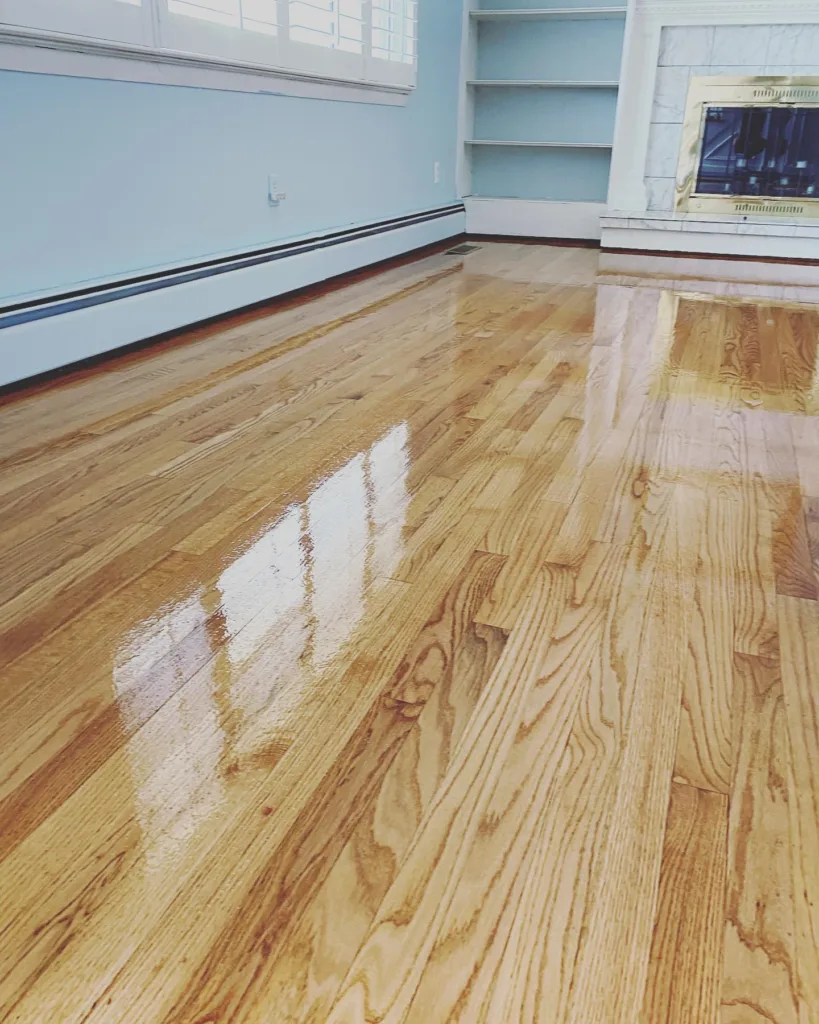 Close-up view of a recently completed hardwood floor refinishing with a high-gloss topcoat, showing a clear window reflection on the surface.